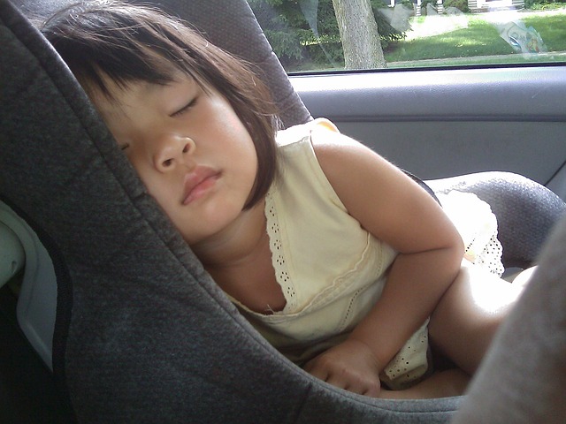 A little girl sleeping in a child's car seat, but she isn't properly strapped in.