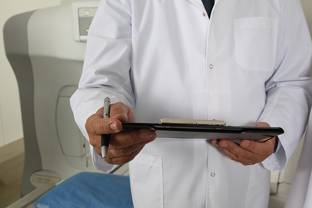 A close up of a doctor, standing in a patient's room and holding a clipboard. They're dressed in a white lab coat.