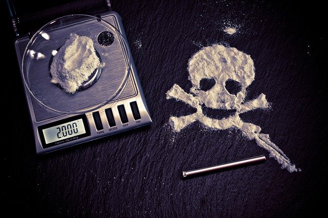 A scale with a portion of white powder on it, and a pile of white powder nearby, made into the shape of a skull, signifying the death that awaits addicts.