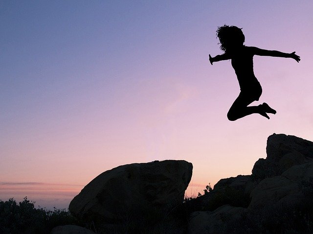 A silhouette of a teen girl jumping off a pile of rocks in front of a sunset sky 