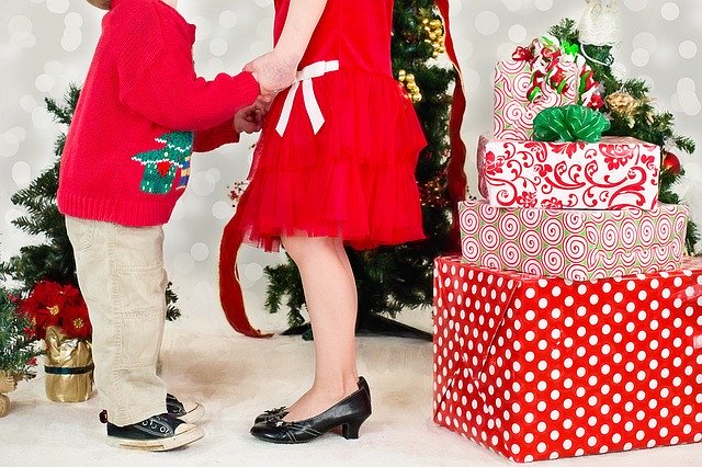 A picture of two children facing each other in front of a Christmas tree. Only their dressy holiday outfits and their feet are visible.