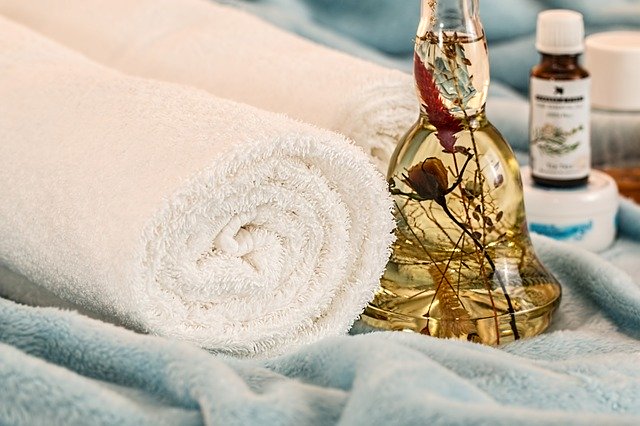 A picture of two white towels rolled up next to a bottle of herb infused massage oil and a blanket.