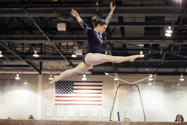 A young female gymnast flies through the air over the bars. In the background you can see the American flagrffe