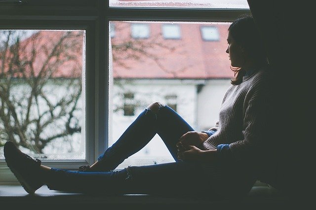 A young woman sitting in a window ledge. She is sitting in darkness and looks sad.